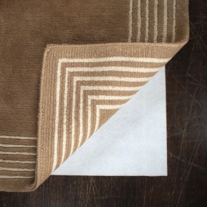May include: A brown rug with white stripes on the edges is folded over a white rug pad. The rug pad is designed to prevent slipping and protect floors.