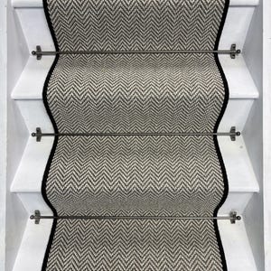 May include: Black and white herringbone patterned carpet runner with black edging, installed on a white staircase with black metal rods and finials.