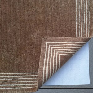 May include: A brown rug with a white border. The rug is folded over to show the underside, which is a white non-slip backing.