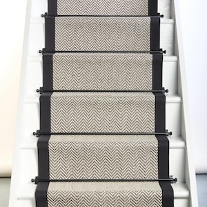 May include: A staircase runner featuring a beige and dark brown herringbone pattern with solid black borders on each side. The runner is secured to the white wooden stairs with horizontal black metal rods placed at the base of each step.