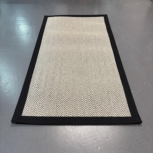 May include: A black and white herringbone patterned rug with a black border.