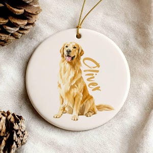 May include: A round, white ceramic ornament featuring a golden retriever illustration. The dog is sitting with its tongue out. The name "Oliver" is written in gold script. A gold string is attached for hanging.
