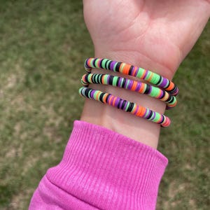 Può includere: Un set di braccialetti colorati a tema Halloween. I braccialetti sono realizzati con piccole perline rotonde e piatte nere, verdi, arancioni e viola. I braccialetti sono impilati su un polso, con una maglia rosa a maniche lunghe sullo sfondo.