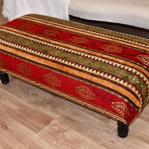 Könnte beinhalten: Gepolsterter Hocker mit einem lebendigen Stoffmuster in Rot, Grün und Gold. Das Design zeigt geometrische Formen und Streifen. Der Hocker hat eine rechteckige Form und steht auf vier schwarzen, gedrechselten Holzbeinen. Länge ca. 90 cm.