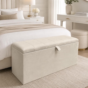 May include: A beige upholstered storage bench with a tufted top and a leather pull tab. The bench is rectangular and sits on a light beige rug. The bedroom setting includes a bed, nightstand, and vanity.