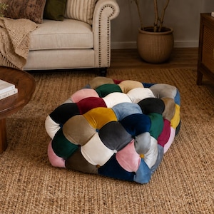 Puede incluir: Un puf de patchwork multicolor con cuadrados de terciopelo en tonos rosa, rojo, azul, verde, amarillo y crema. Descansa sobre una alfombra de yute tejida. En el fondo se ven un sofá neutro y muebles de madera.