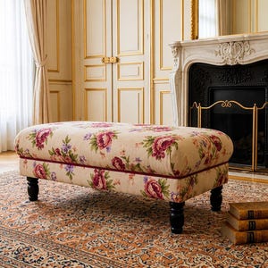 May include: An upholstered ottoman with a floral pattern in pink, purple, and green on a cream background. The ottoman has a dark red trim and sits on four black legs. The room features a fireplace, a patterned rug, and a stack of books.