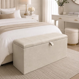 May include: A beige upholstered storage bench with a tufted lid sits at the foot of a bed. The bedroom features a neutral color palette, with a white bedspread, a beige rug, and a white nightstand with a lamp.