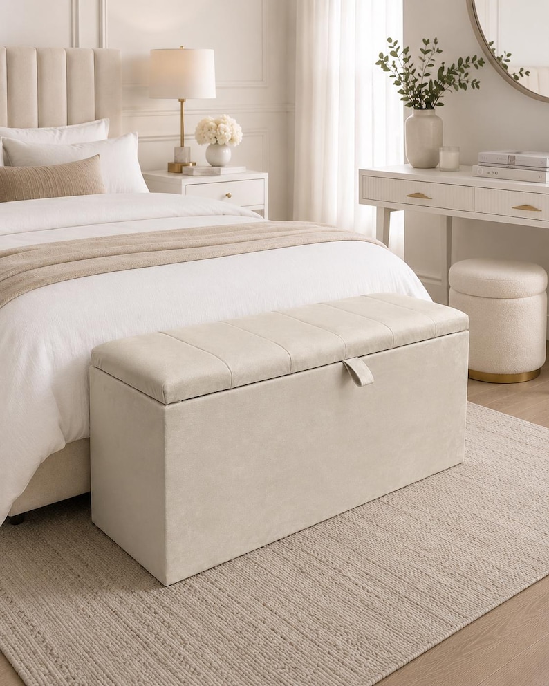 May include: A beige upholstered storage bench with a tufted lid sits at the foot of a bed. The bedroom features a neutral color palette, with a white bedspread, a beige rug, and a white nightstand with a lamp.