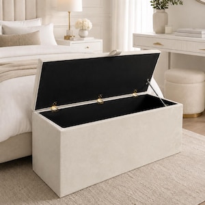 May include: A beige rectangular storage ottoman with a hinged lid, open to reveal a black interior. The ottoman is placed on a neutral-toned rug in a bedroom setting. Gold-colored hinges and supports are visible.