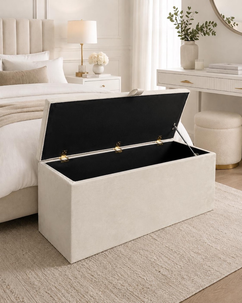 May include: A beige rectangular storage ottoman with a hinged lid, open to reveal a black interior. The ottoman is placed on a neutral-toned rug in a bedroom setting. Gold-colored hinges and supports are visible.