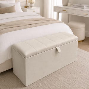 May include: A beige rectangular storage bench with a tufted lid sits at the foot of a bed. The bench has a rectangular shape and a neutral color, blending with the bedroom's decor. The room features a neutral color palette with a rug and a white nightstand.