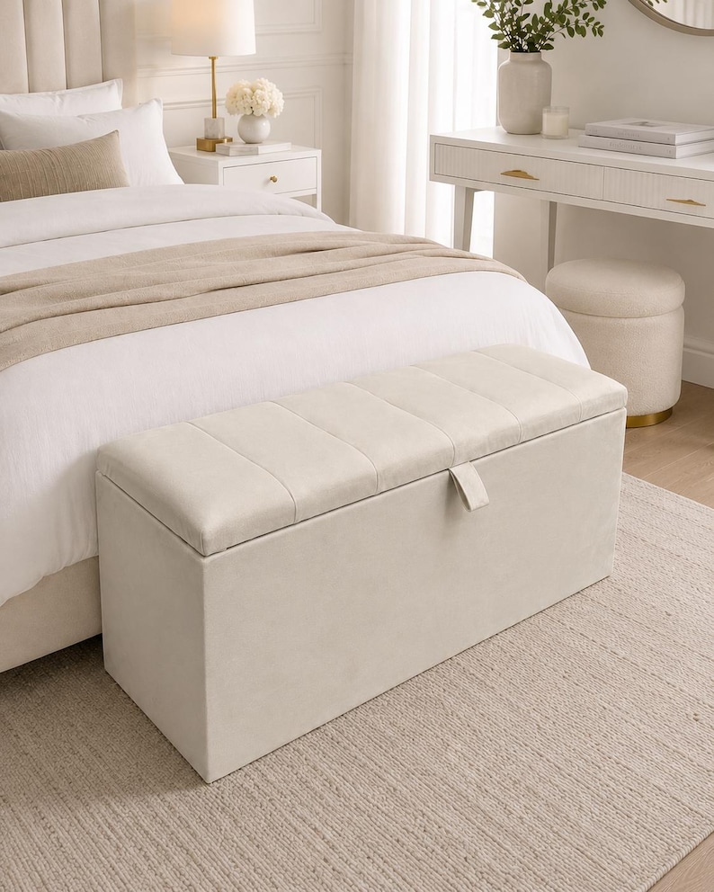 May include: A beige rectangular storage bench with a tufted lid sits at the foot of a bed. The bench has a rectangular shape and a neutral color, blending with the bedroom's decor. The room features a neutral color palette with a rug and a white nightstand.