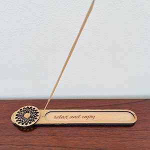 Puede incluir: Porta incienso de madera con un diseño de corte circular y el texto "relax and enjoy" grabado en la superficie. El porta incienso es marrón y tiene un acabado de madera natural.