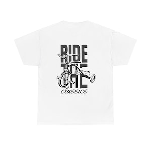 Può includere: T-shirt bianca con grafica nera di un triciclo e la scritta "RIDE THE classics". Il design è centrato, presentando uno stile classico e retrò. La maglietta è realizzata in materiale morbido.