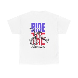 Può includere: T-shirt bianco con un'illustrazione in bianco e nero di un triciclo classico. Il testo "RIDE THE classics" è stampato sopra il triciclo in lettere a blocchi rosse e blu. La maglietta è realizzata in un materiale morbido e confortevole.