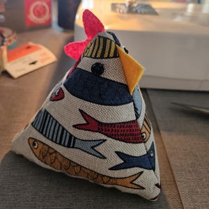 May include: A handmade fabric chicken-shaped doorstop. The doorstop features a colorful design with fish and stripes on a cream background. It has a red comb, yellow beak, and a black button eye. The doorstop is likely used for home decor.