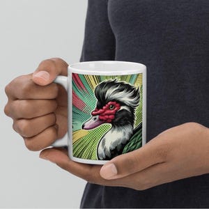Peut inclure: Une tasse en céramique blanche avec une illustration colorée d'un canard avec une crête. Le canard a un bec rouge et un corps vert et noir. L'arrière-plan est un design de style bande dessinée coloré.