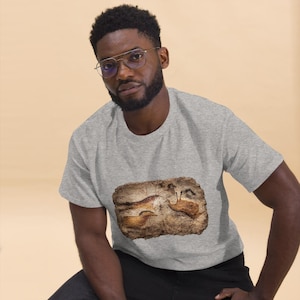 Op de afbeelding: Een grijs t-shirt met een gedetailleerde afbeelding van oude grottekeningen in bruin- en beigetinten. Het kunstwerk toont dierenfiguren en is gecentreerd op het shirt.