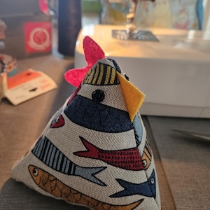 May include: A pyramid-shaped fabric chicken ornament featuring a cream-colored background with a repeating pattern of blue, red, and yellow fish. The chicken has a bright red felt comb, a yellow felt beak, and a small black button for an eye.