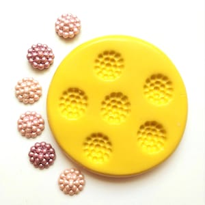 May include: A yellow silicone mould with six flower-shaped impressions. Several small, round, pearl-like embellishments in shades of pink and mauve are scattered around the mould, suggesting a crafting or jewellery-making theme.