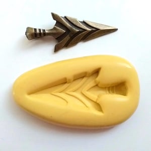 May include: A bronze arrowhead and a yellow silicone mould in the shape of an arrowhead. The arrowhead has a detailed design with a textured surface. The mould is oval-shaped.