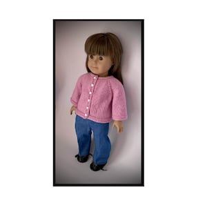 May include: A pink knitted cardigan with white buttons, worn by a doll with brown hair and blue jeans.