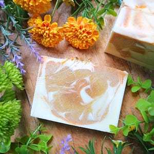 Peut inclure: Savons artisanaux avec un motif tourbillonnant blanc et orange, présentés avec des fleurs orange vif et du feuillage sur une surface en bois. Le savon a un aspect marbré, suggérant des ingrédients naturels.