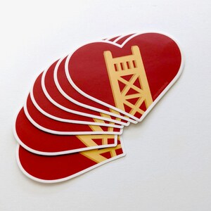 May include: A stack of red heart-shaped stickers with a gold outline. Each sticker features a stylized illustration of a bridge.