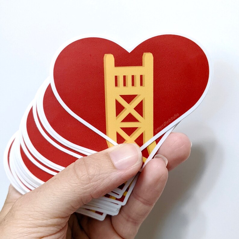 May include: A stack of red heart-shaped stickers with a yellow graphic of a bridge inside. The sticker has the text "@mycraftedstudio" on the bottom right corner.