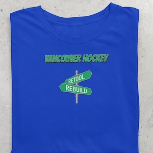 May include: Royal blue t-shirt with the text "Vancouver Hockey" in green above a green street sign graphic. The street sign reads "RETOOL" and "REBUILD".