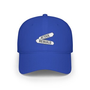 May include: A royal blue baseball cap with a curved brim. The cap features a white and gray emblem with the words "RETOOL" and "REBUILD" in a road sign design. The cap is made of a soft material.