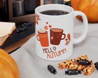 May include: White ceramic mug with a "Hello Autumn" design featuring two coffee cups, hearts, and fall leaves. The mug is filled with a dark liquid and is surrounded by Halloween candy and pumpkins.