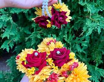 May include: Floral wrist corsages featuring yellow chrysanthemums, deep red roses, and small yellow and red accent flowers. The corsages are adorned with white pearl bracelets. The background is a lush green foliage.
