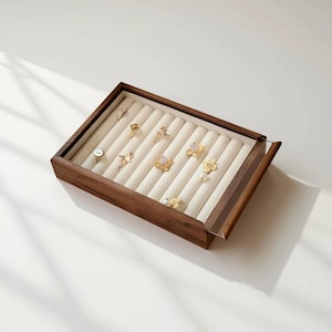 May include: A wooden jewellery box with a clear lid, displaying a collection of rings. The rings have diverse designs, including floral, gemstone, and pearl details. The box has a light-coloured, cushioned interior.