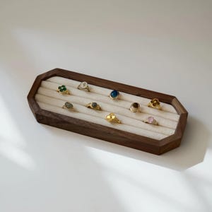 Puede incluir: Una bandeja de joyería de madera oscura con interior crema muestra varios anillos. Los anillos presentan diferentes piedras preciosas y diseños, incluyendo una flor, un trébol y una piedra redonda.