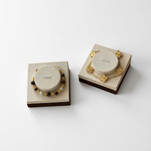 Op de afbeelding: Twee sieradendisplays tonen armbanden. Eén heeft bruine en witte kralen met gouden accenten. De andere toont een gouden klavervormige armband. Beide worden gepresenteerd op beige platforms met bruine randen, tegen een witte achtergrond.