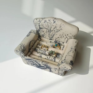 Puede incluir: Un joyero pequeño, tapizado en forma de sillón, con tela color crema con un diseño de árboles y follaje azul. La caja abierta muestra varios anillos con bandas doradas y piedras preciosas de colores.