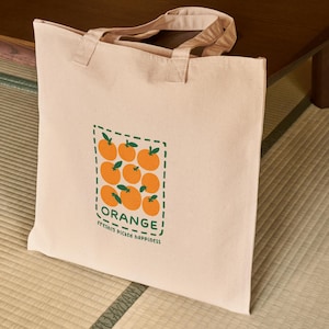 May include: A beige canvas tote bag featuring a rectangular graphic of orange fruits with green leaves. The word "ORANGE!" is printed above the fruits, with "freshly picked happiness" below. The bag has matching beige handles.
