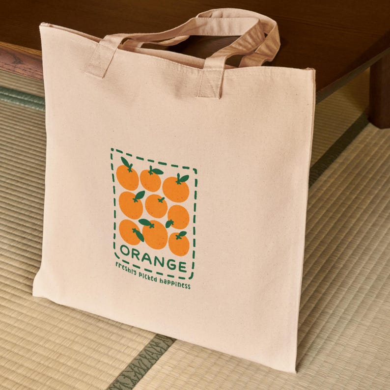May include: A beige canvas tote bag featuring a rectangular graphic of orange fruits with green leaves. The word "ORANGE!" is printed above the fruits, with "freshly picked happiness" below. The bag has matching beige handles.
