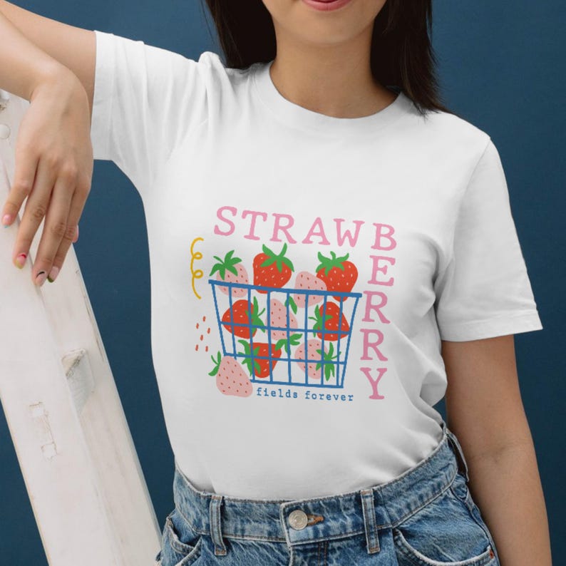 May include: White t-shirt featuring a colorful graphic of strawberries in a blue basket with the text "STRAWBERRY fields forever" in pink and white. The design includes red, pink, and green elements, creating a playful and summery vibe.