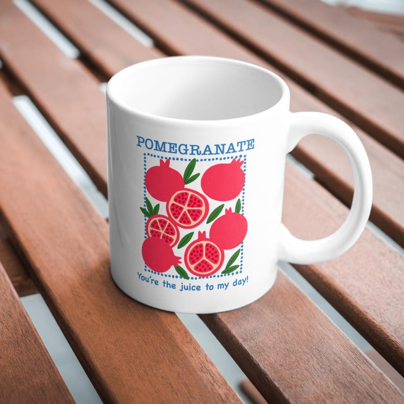 May include: White ceramic mug featuring a pomegranate design with the word "Pomegranate" in blue. The design includes whole and sliced pomegranates with green leaves. The text "You're the juice to my day!" is also included.
