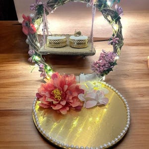 Può includere: Un portatore di anelli nuziali decorativo con una base dorata e una ghirlanda floreale. La ghirlanda sostiene una piccola altalena che contiene due scatole per anelli. La base è adornata da un bordo di perle e un grande fiore rosa.