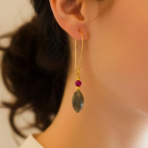 May include: Gold-tone drop earrings with a ruby-colored bead and a gray-green, oval-shaped stone. The earrings are suspended from a curved wire and are worn on the ear. The earrings are a piece of jewelry.