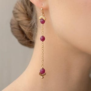 Puede incluir: Pendientes colgantes dorados con un diseño geométrico y tres piedras preciosas de color rubí. Los pendientes cuelgan de un gancho y están conectados por una delicada cadena. Un accesorio de joyería.
