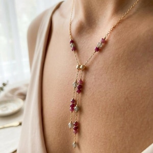 May include: A gold chain lariat necklace featuring small, faceted, round beads in shades of deep pink and translucent grey. The necklace has a central gold bead connector from which two delicate chains dangle, each accented with alternating pink and grey beads.