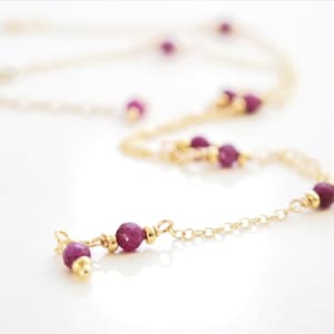 May include: A delicate gold necklace with small, faceted ruby beads. The necklace features a fine chain and is adorned with several ruby accents, creating a simple yet elegant design. The ruby beads are a deep, rich red color.