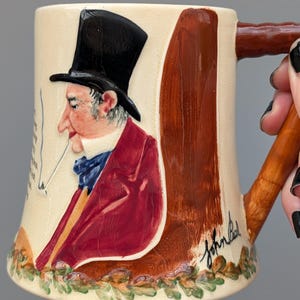 May include: Cream ceramic mug with a relief portrait of a man in a top hat and red coat, smoking a cigarette. The mug has a brown handle and base with a painted design. The artist's signature is visible.