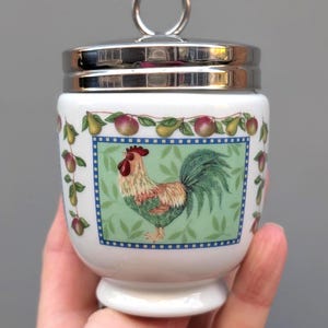 May include: White ceramic egg cup with a silver lid and handle. The cup features a colourful rooster illustration on a green background, framed with a blue border. The design includes fruit and foliage.