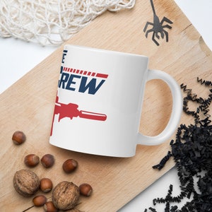 Pode incluir: Caneca de cerâmica branca com o texto "The Crew" em azul e vermelho, e um gráfico vermelho de motosserra. A caneca está sobre uma tábua de madeira com nozes e decorações de Halloween.